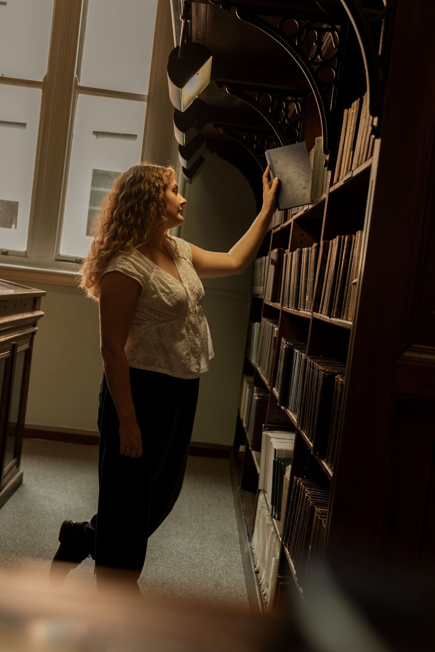 Figure reaching for books on tall library shelves in atmospheric lighting. Cinematic Photography Scenes in Wigan
