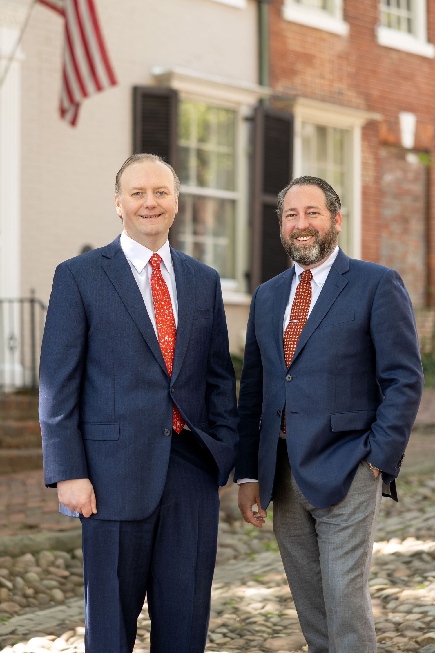 Two professionals in navy suits with orange ties standing together outside a brick building with an American flag.