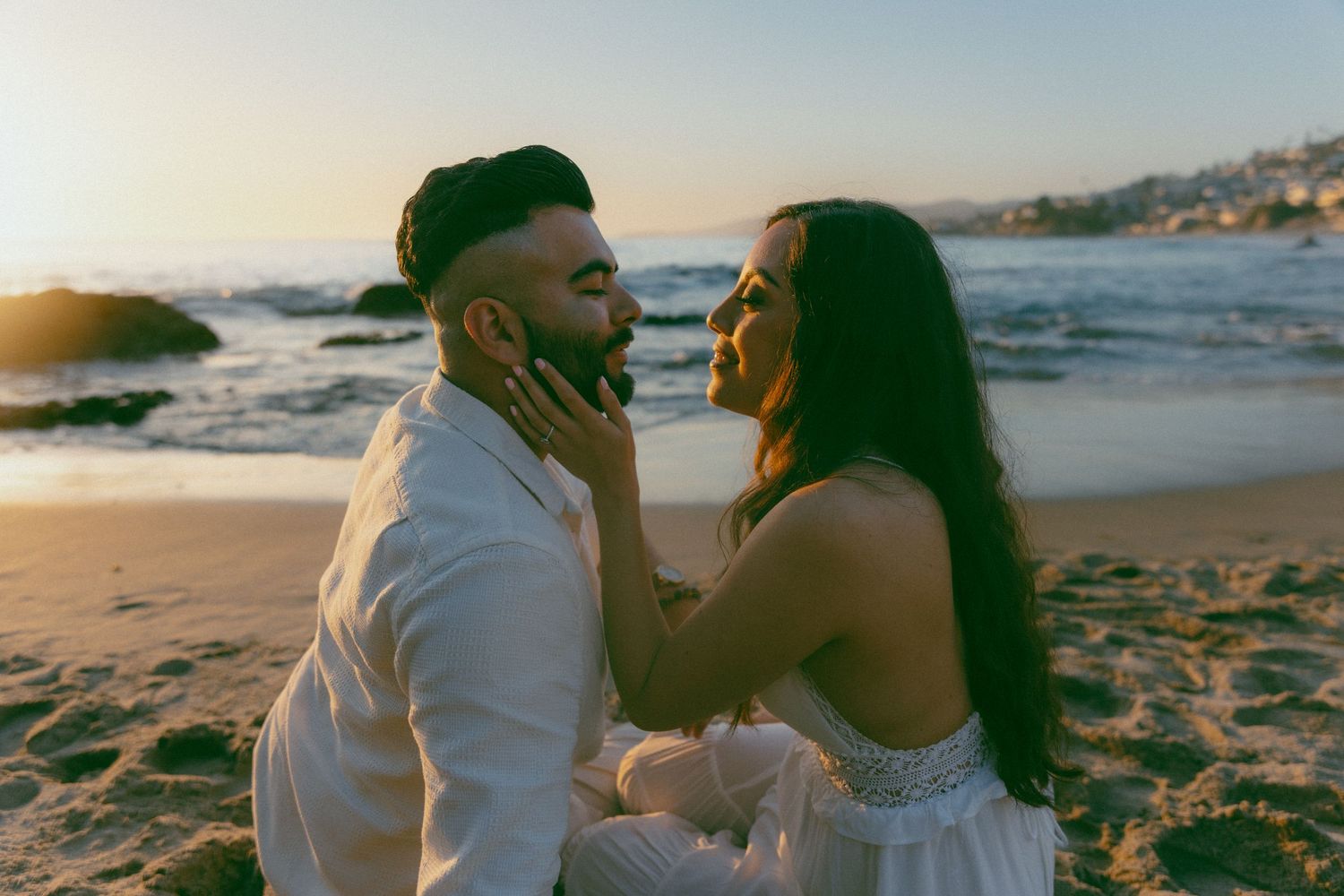 A romantic couple shares an intimate moment on a sandy beach at sunset along the coastline.