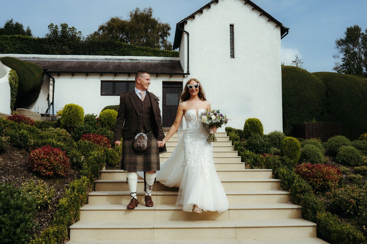Bride and groom in Scottish kilt holding hands on steps outside white wedding venue surrounded by manicured hedges.
