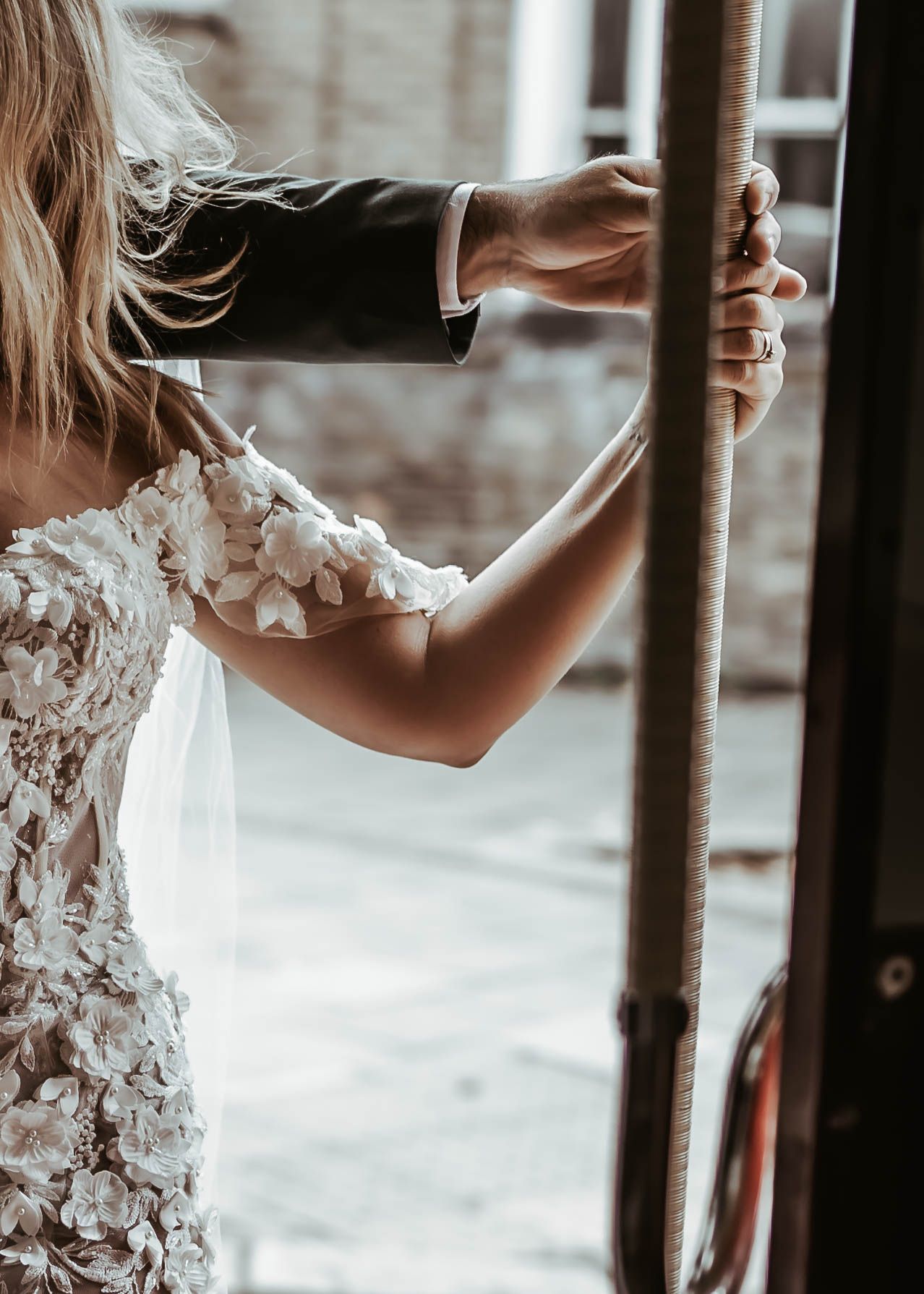 Close-up view of a bride in white lace dress holding onto a railing with sunlit window in background.