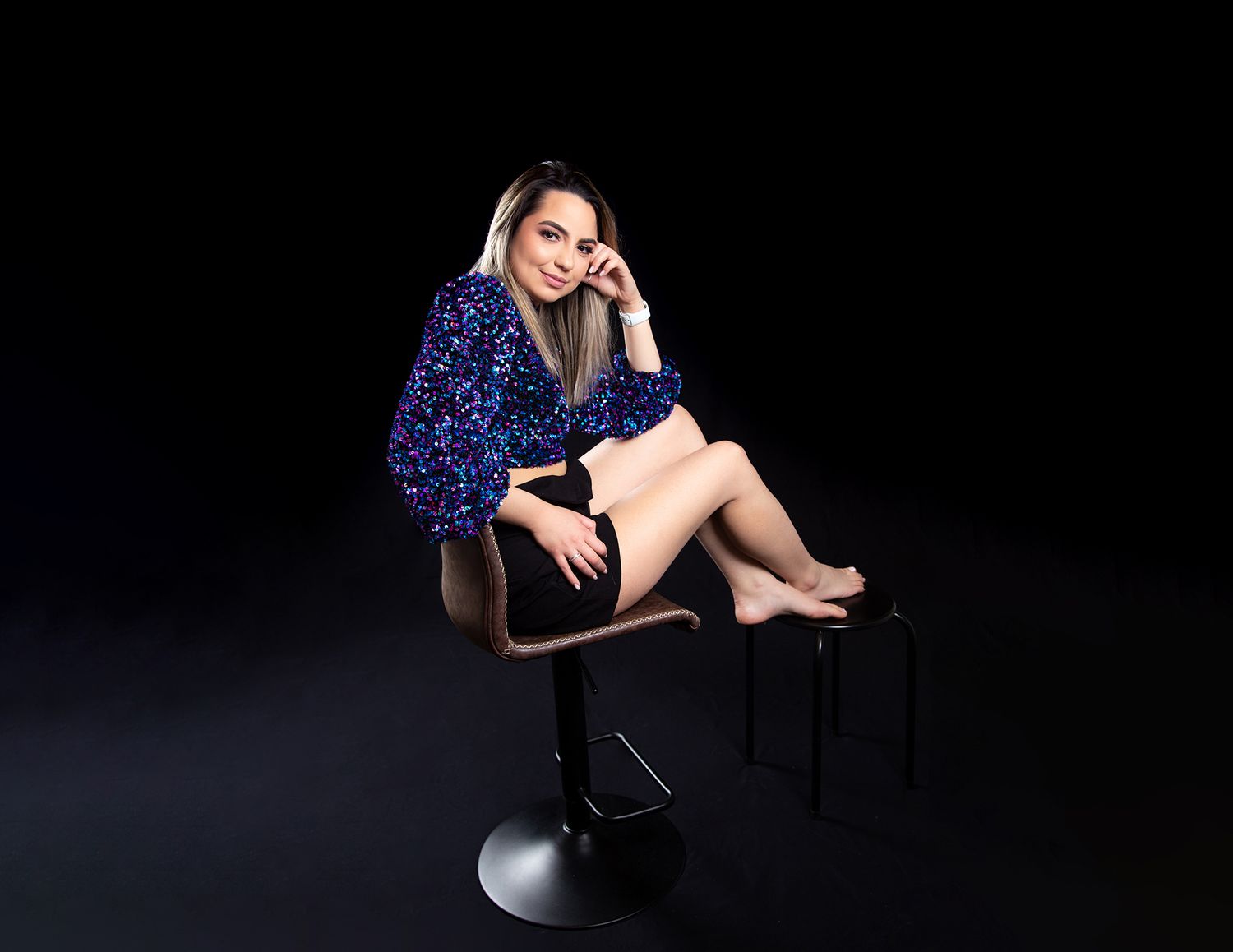 A person in a sparkly blue sequin jacket sits barefoot on a black bar stool against a dark background.