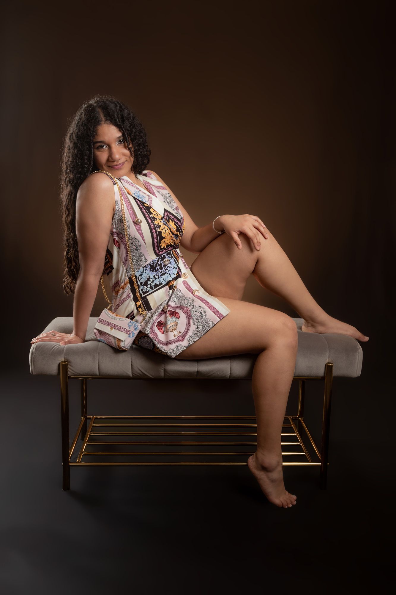 Woman in patterned dress poses on leather bench against dark brown background in studio setting.