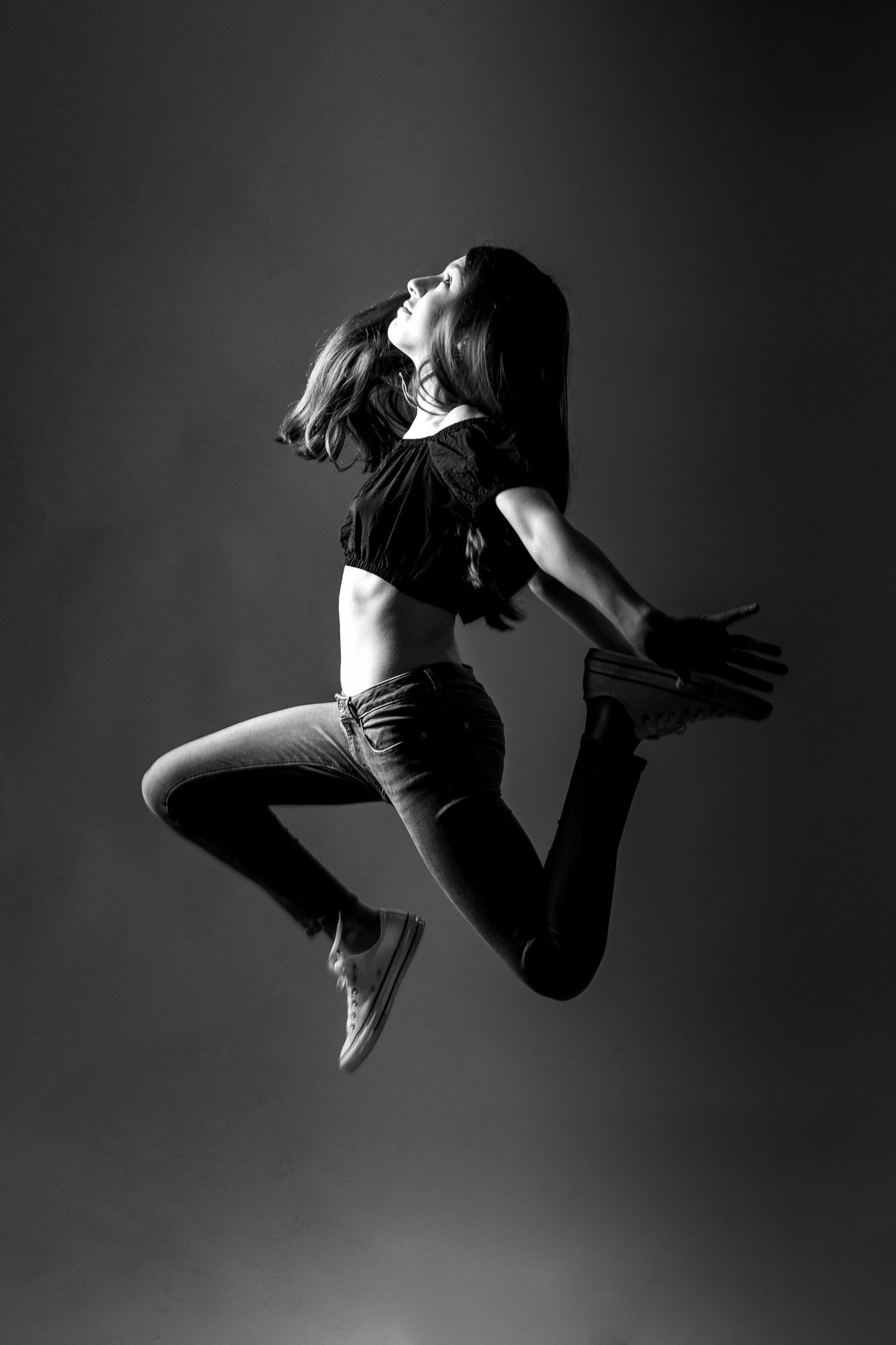 Dramatic black and white photo of a dancer leaping in mid-air wearing casual clothes against dark background.