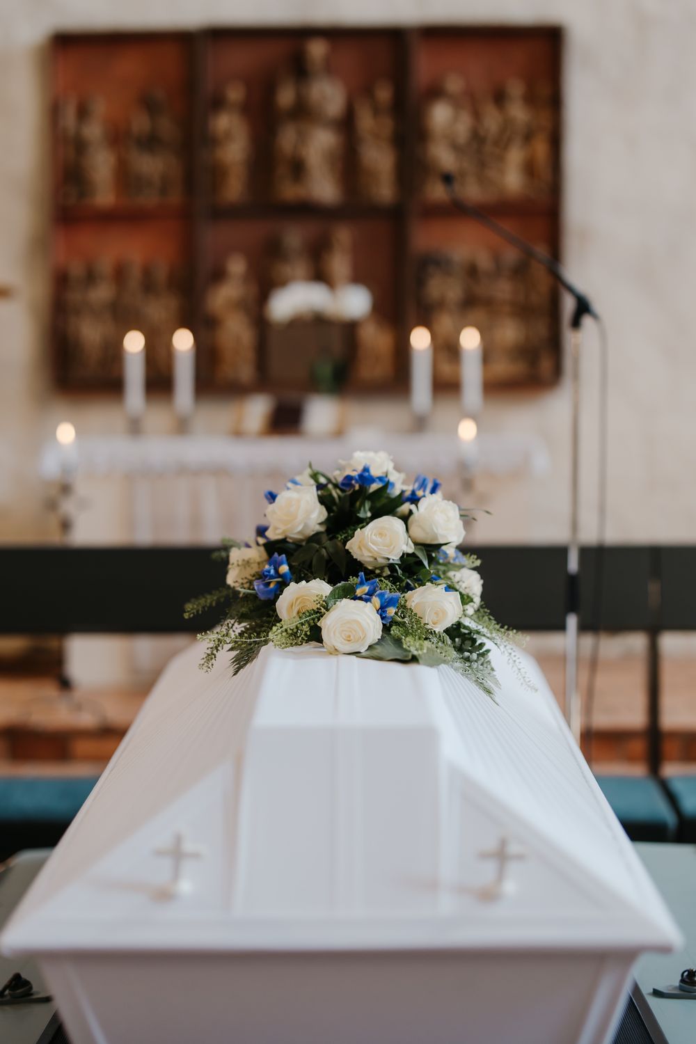 White casket with white and blue floral arrangement in a church setting with candles and altar in background.