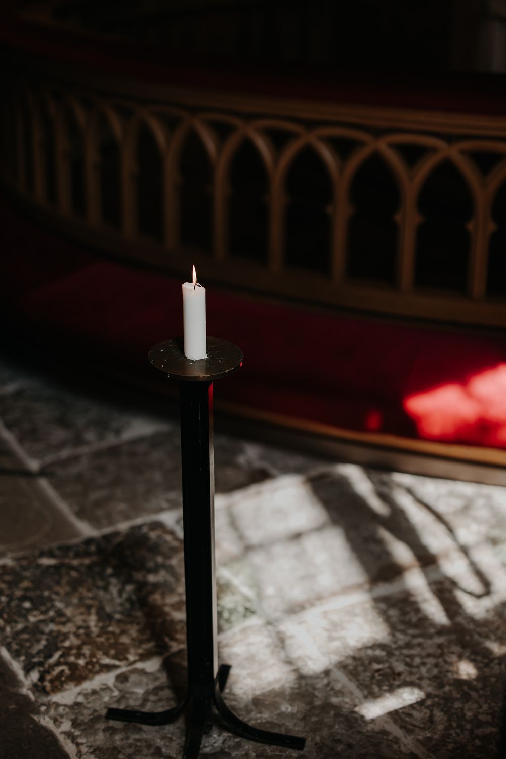 A single white candle on a black iron stand in a dimly lit church with gothic arched woodwork in background.