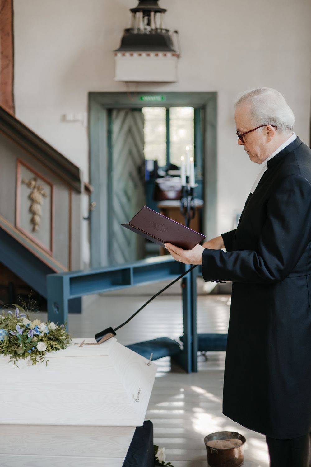 A minister in black robes stands at a white altar in a sunlit church.