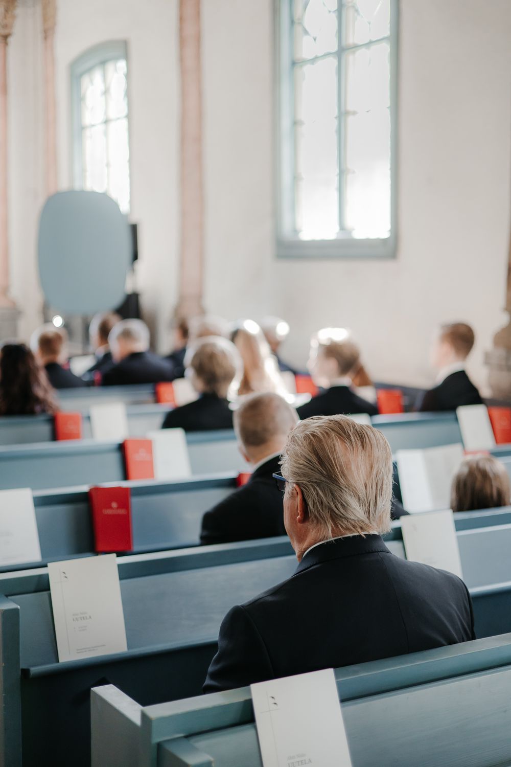 People sitting in rows of white church pews during a religious service in a bright, sunlit sanctuary.