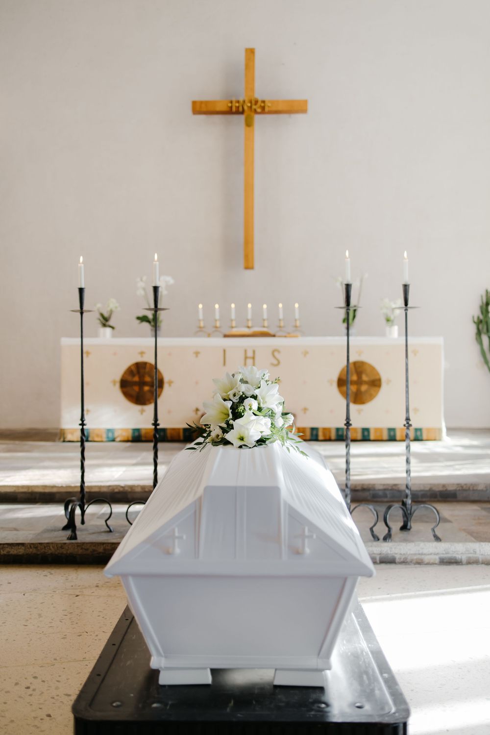 White coffin adorned with candles and wooden cross in a simple, serene church setting.