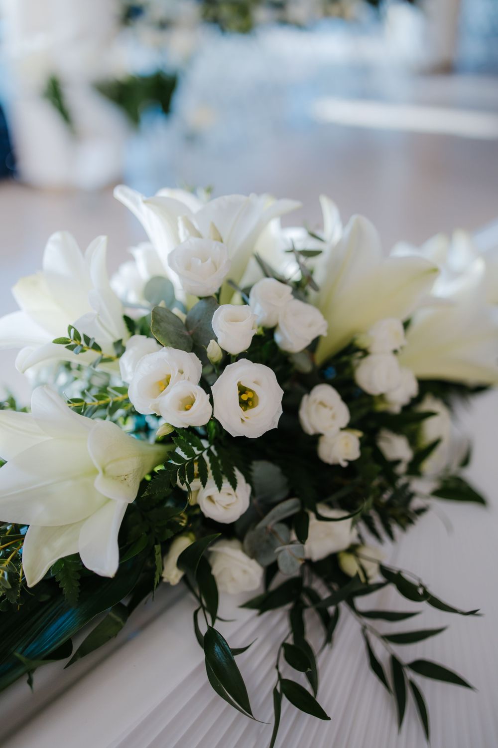 Elegant white floral wedding centerpiece arrangement with lilies and greenery on a light tablecloth.
