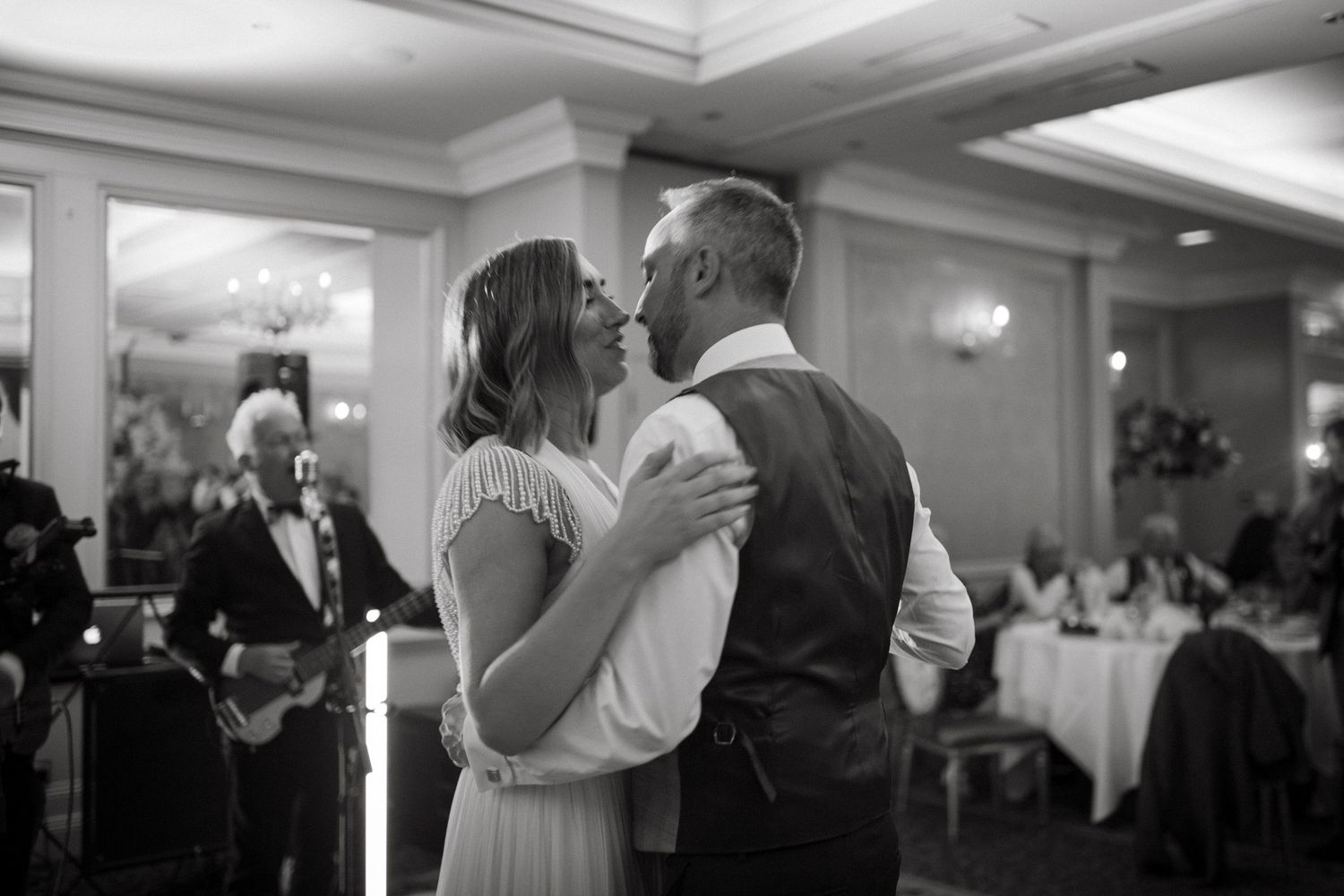 A couple shares their first dance at an elegant wedding reception in a ballroom with chandeliers and tables in the background.