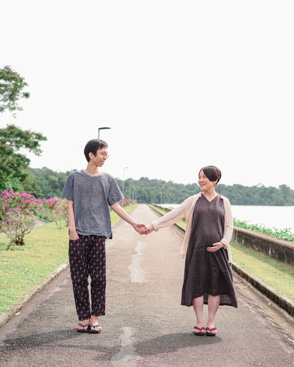 Two people in casual attire hold hands while standing on a scenic path with greenery and a lake in the background.