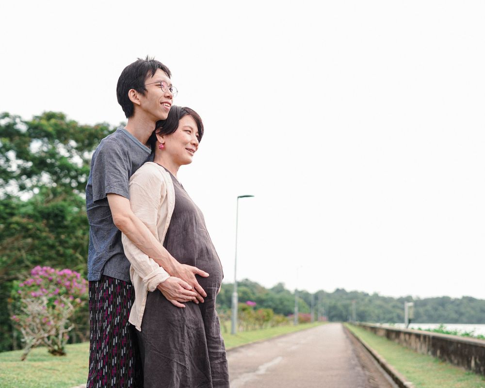 A couple embraces while standing on a peaceful path with green trees and nature in the background.