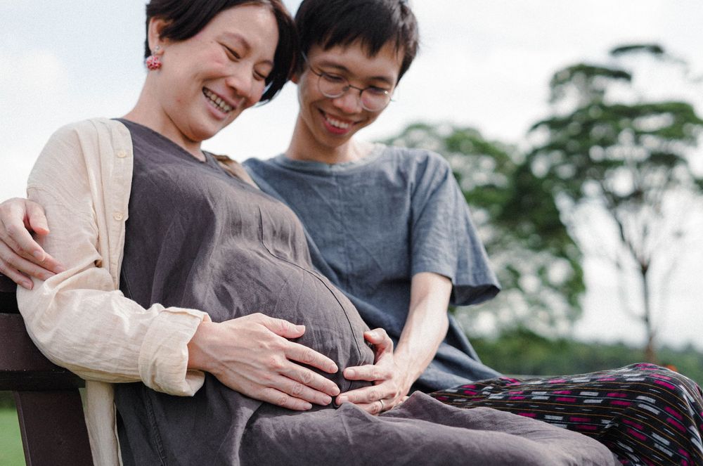 A couple shares a tender moment outside while expecting a baby, with green foliage in the background.
