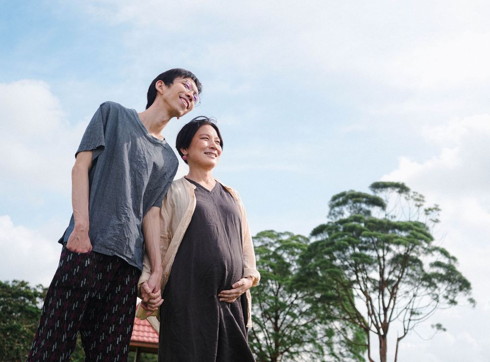 A couple stands together against a blue sky backdrop and lush green trees while holding hands.