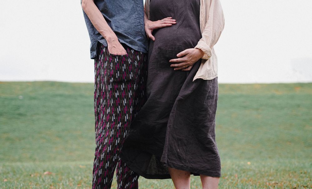 Standing pregnant couple in casual clothing embrace on grassy field against light background.