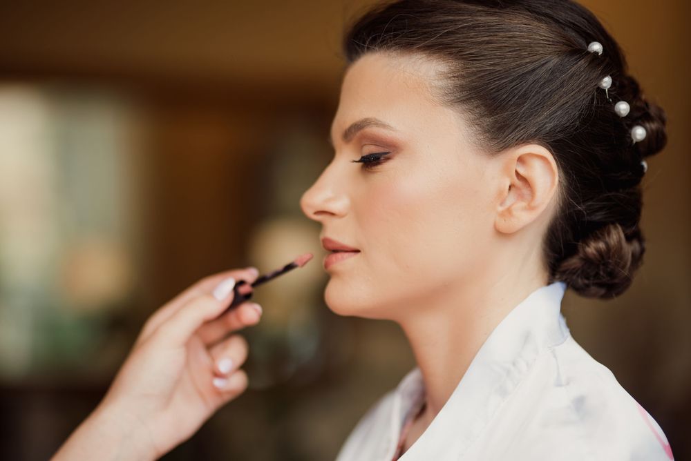 Makeup artist applying lip color to bride wearing white silk robe during wedding preparation.