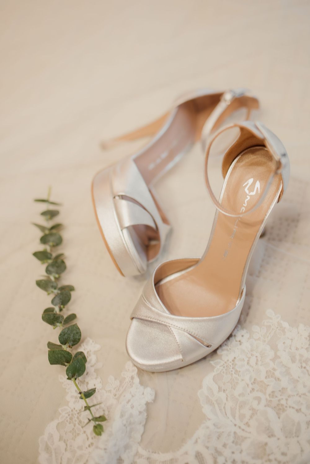 Elegant silver wedding heels with delicate straps displayed with eucalyptus and lace.
