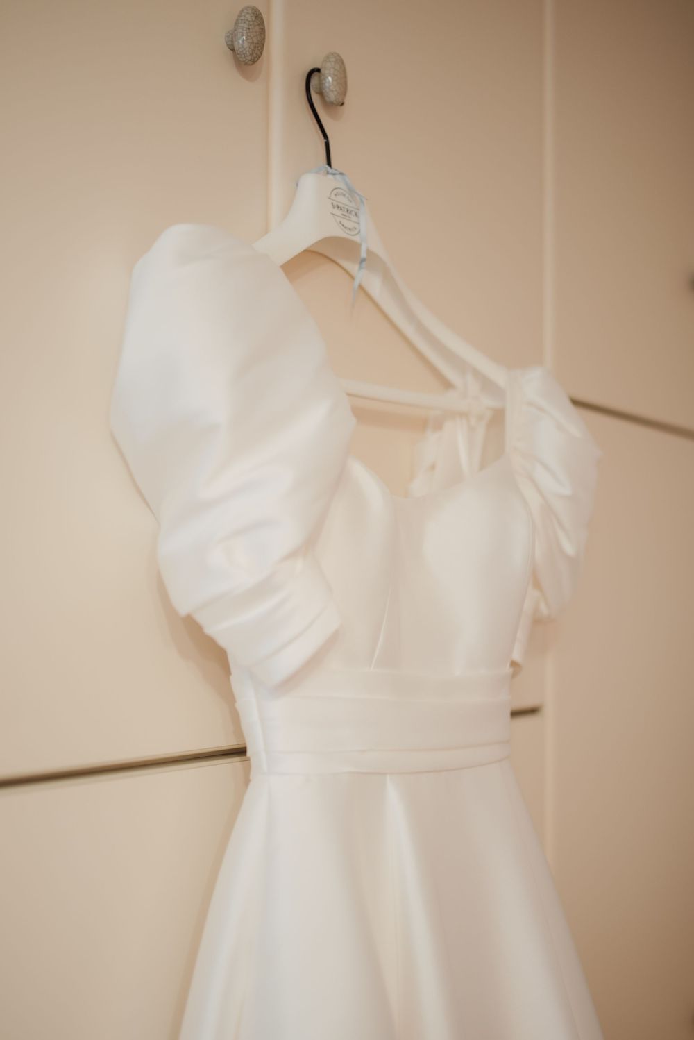White wedding dress with ruffled details hanging on padded hanger against neutral wall.