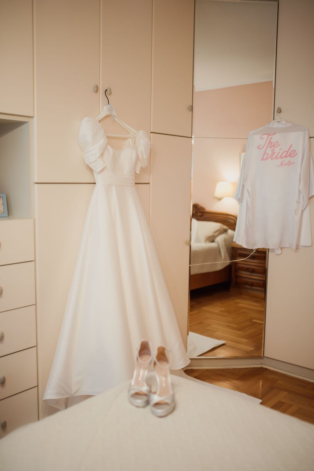 A classic white wedding dress hangs on display with matching heels and a bridal robe in a dressing room.
