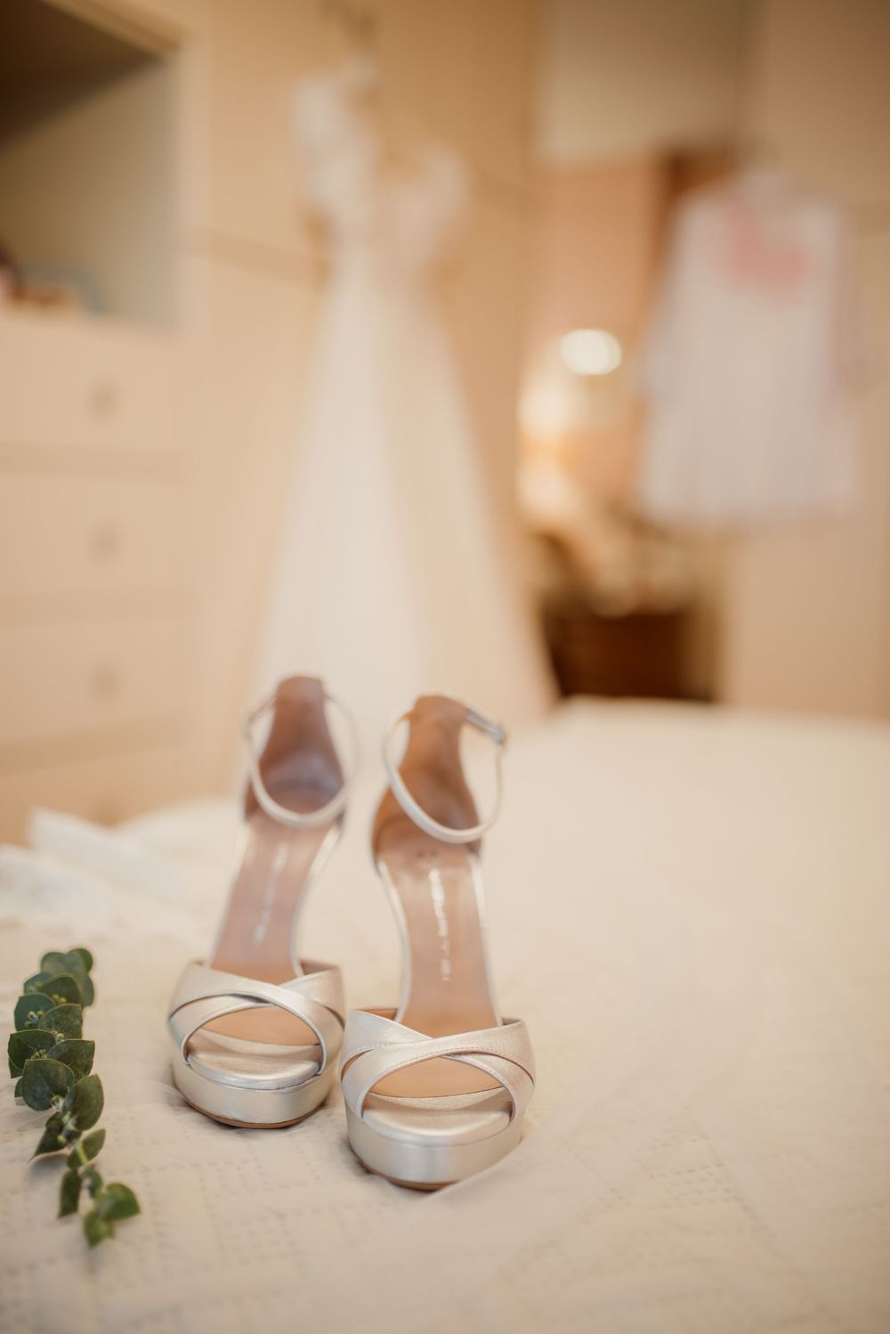 Silver bridal shoes with delicate straps displayed next to wedding rings and greenery.