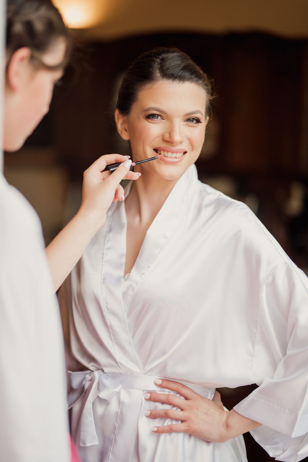 Smiling woman in white satin robe getting professional makeup done during bridal preparation.