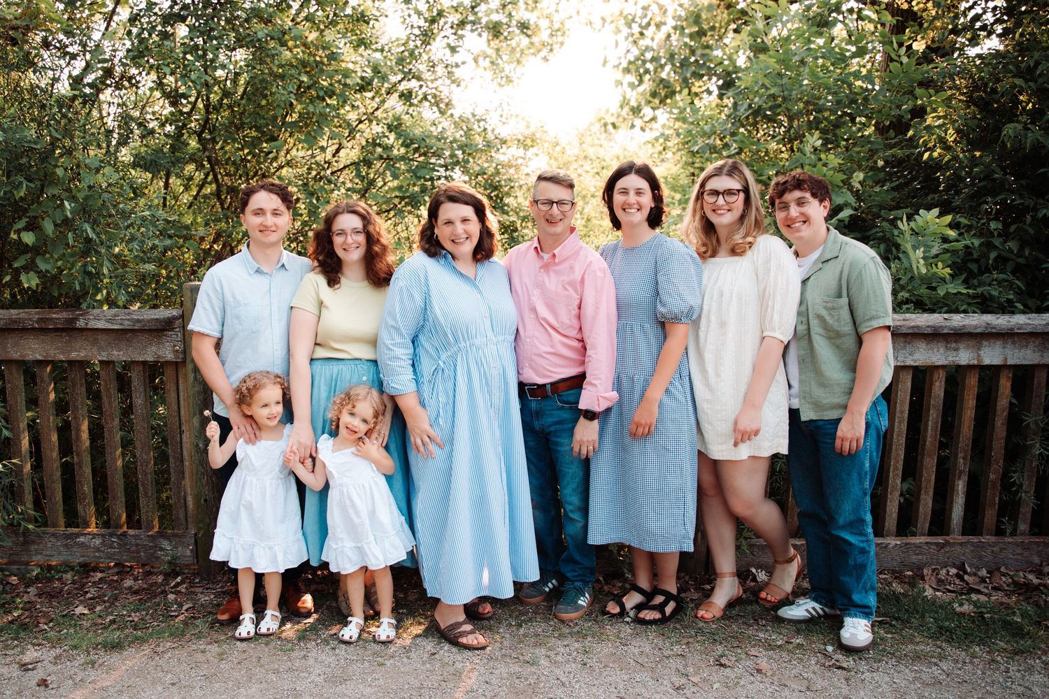 Extended family group in coordinated blue and pastel outfits poses on wooden deck surrounded by trees.