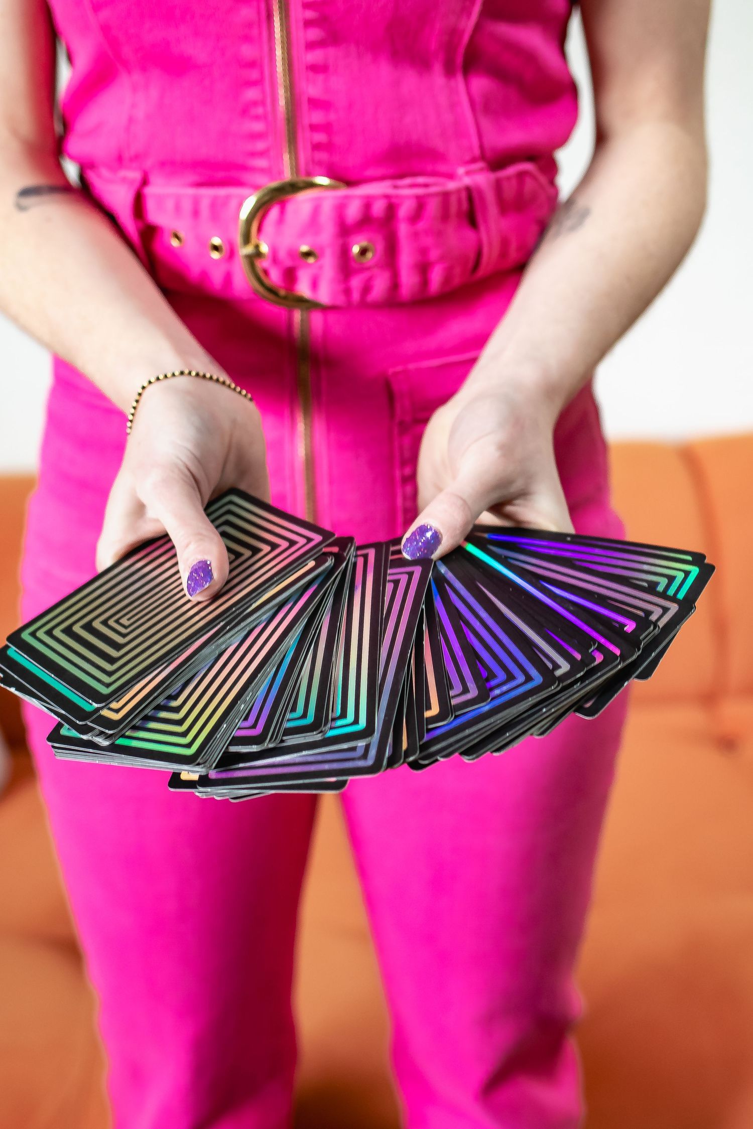 Hands holding fanned out holographic tarot cards while wearing a bright pink outfit and purple nail polish.
