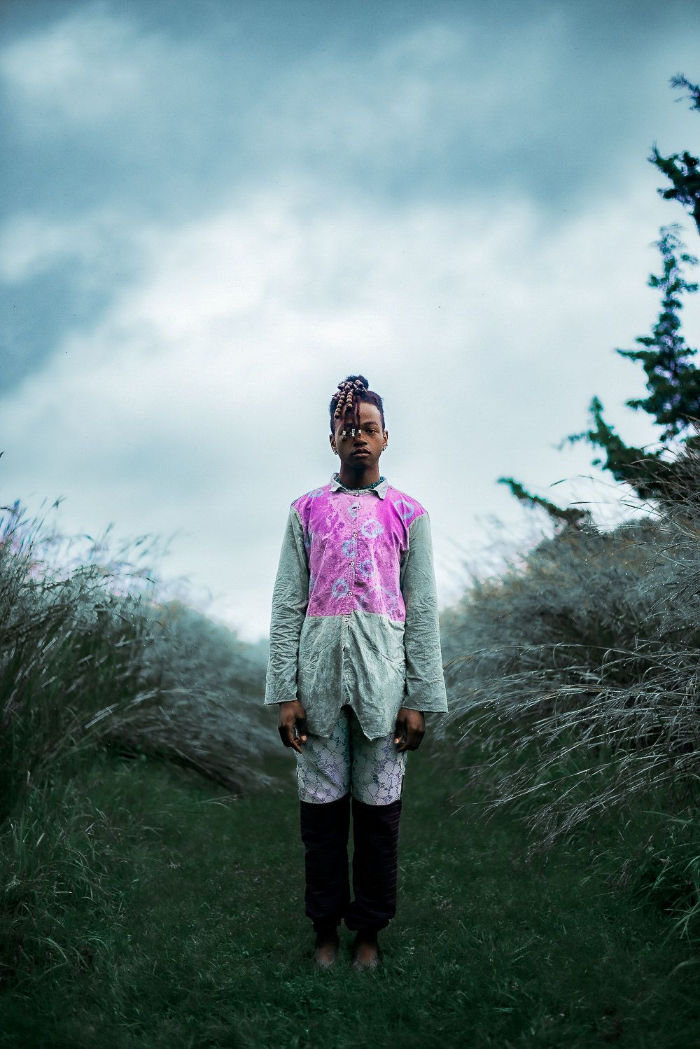 A person wearing a pink and gray jacket stands in tall grass against a moody cloudy sky in a surreal outdoor scene.