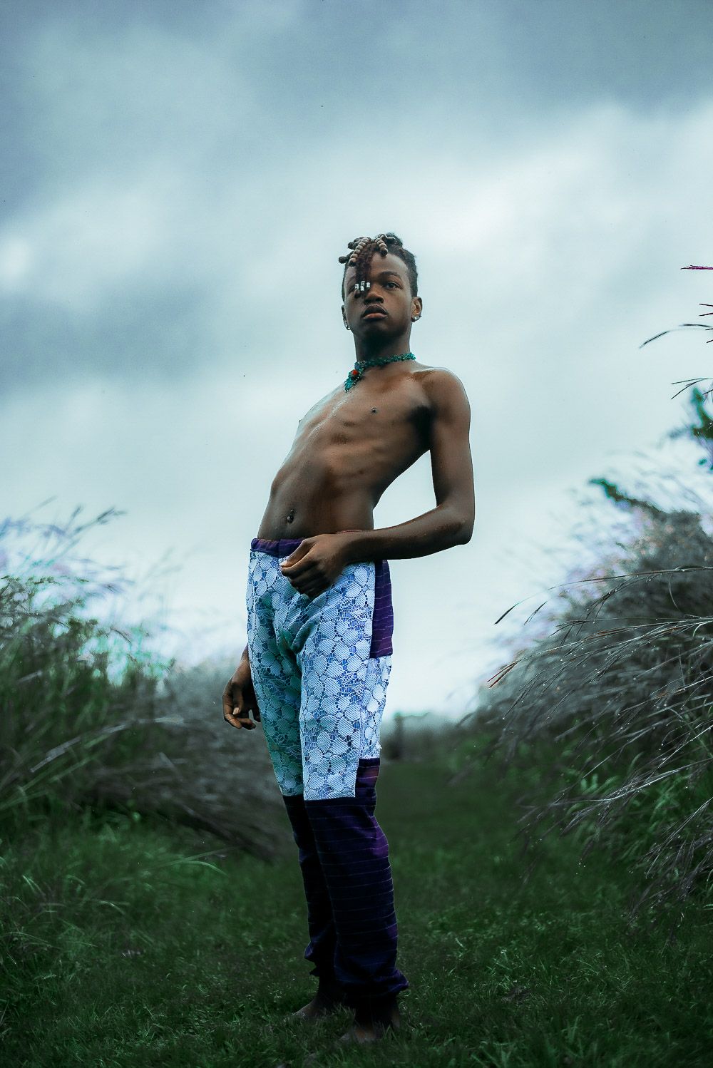 A shirtless figure stands in tall grass against a moody sky wearing patterned shorts in a dramatic nature portrait.