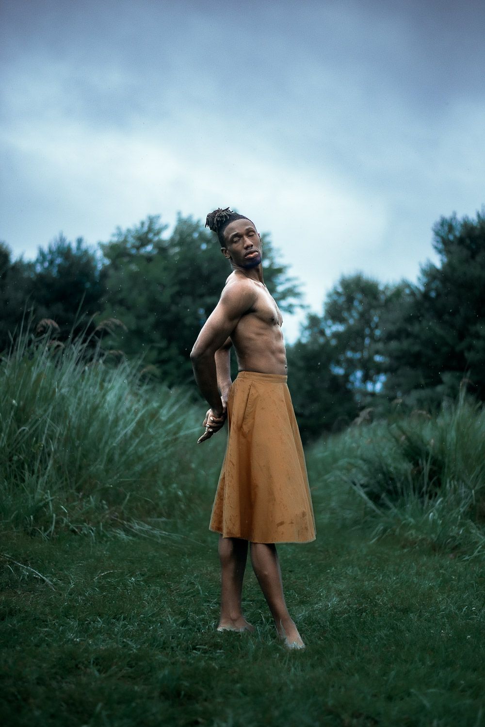 A figure stands barefoot in tall grass wearing an orange skirt against a backdrop of pine trees on a cloudy day.
