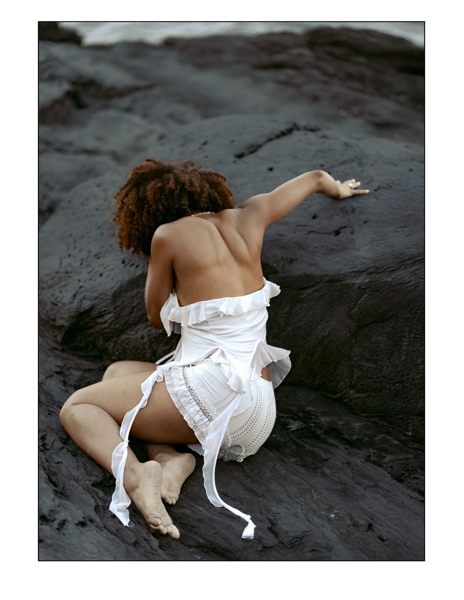 Figure in a white flowing dress poses artistically on dark volcanic rocks near the ocean.