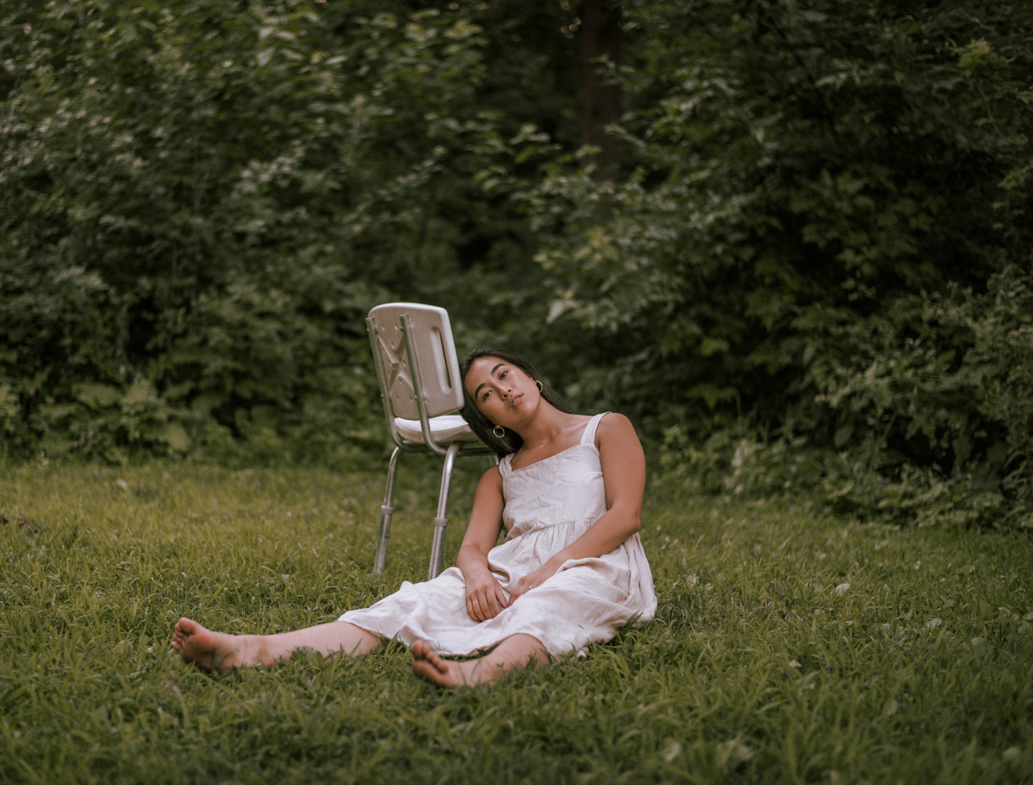 A person in a white dress reclines against a chair in a sunlit meadow.