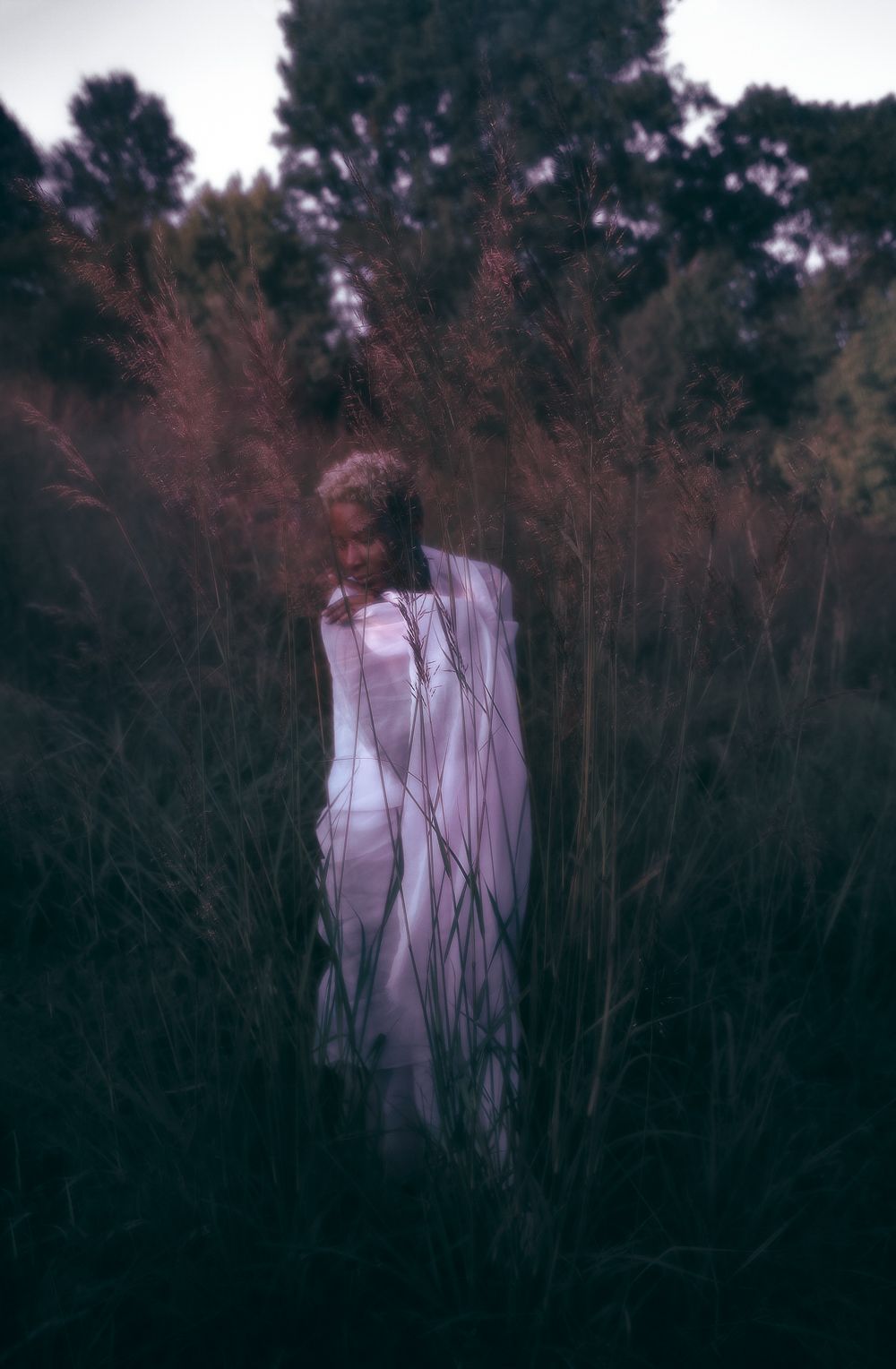 Ethereal figure in white dress stands among tall grass at dusk creating a haunting silhouette against darkening sky.