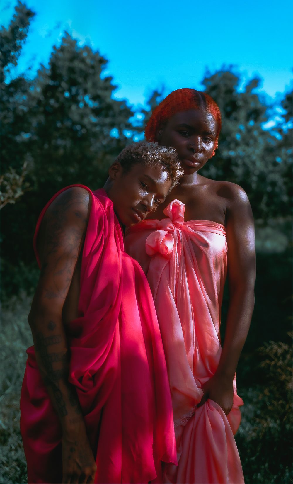 Two figures in flowing pink and red dresses pose together outdoors at dusk against a backdrop of trees.