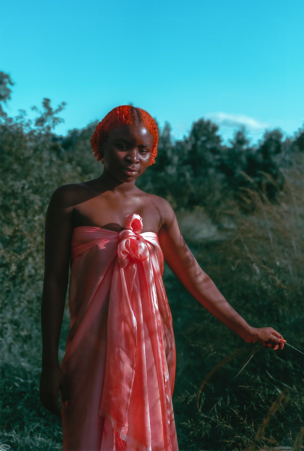 A figure in a flowing coral dress stands gracefully in a natural outdoor setting during golden hour.