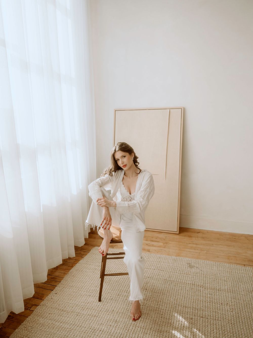 A woman in a white linen ensemble sits elegantly on a wooden stool near sheer curtains and a neutral area rug.