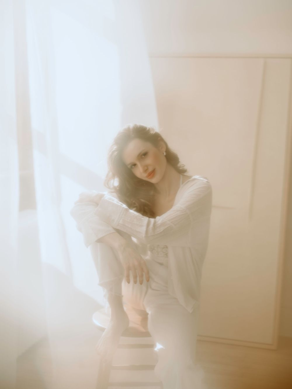 Ethereal portrait in soft white tones shows a dreamy scene with sheer curtains and natural window light creating a gentle atmosphere.