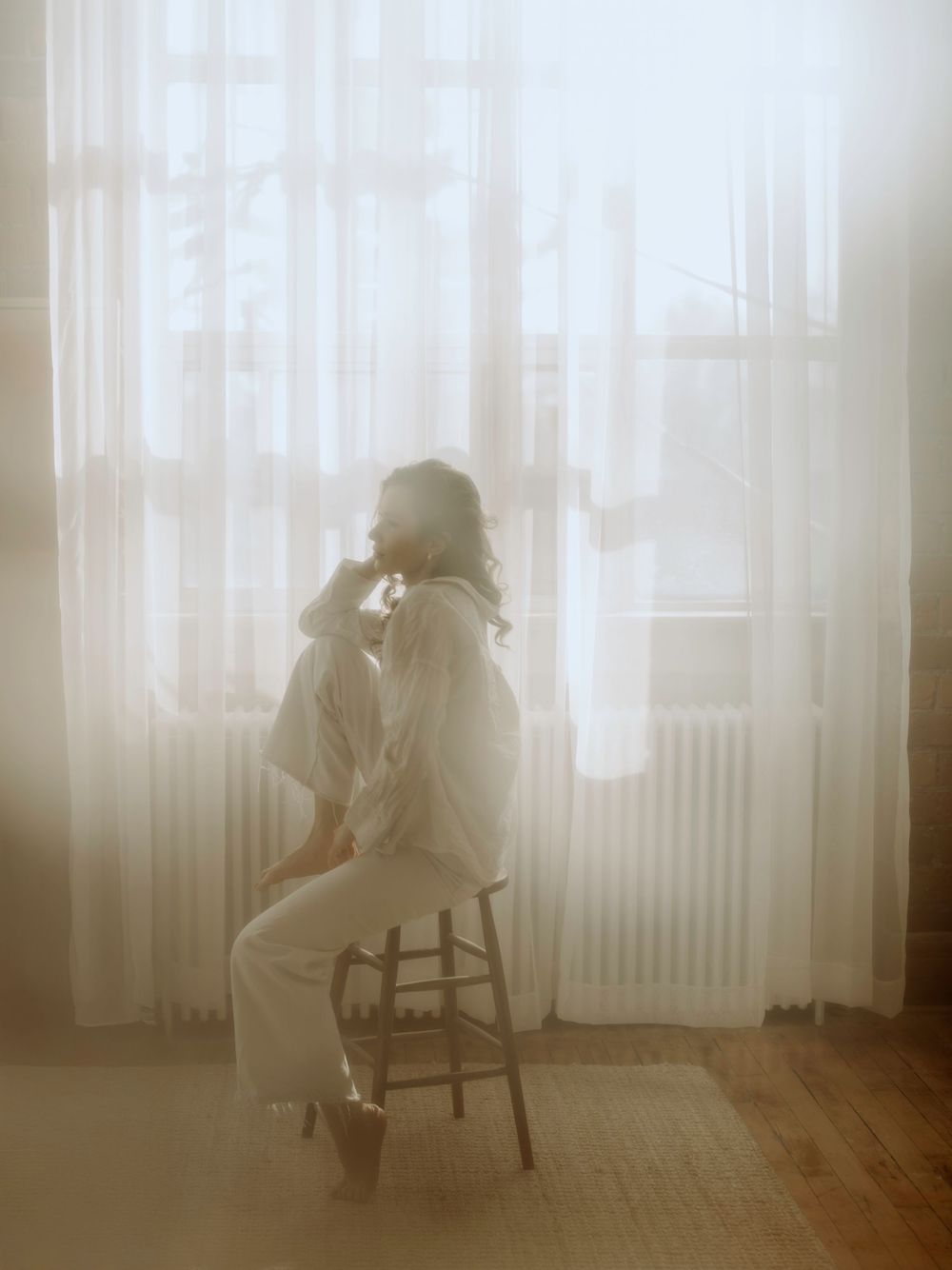 A silhouette figure sits on a wooden chair by a window with sheer curtains in a dreamy, ethereal atmosphere.