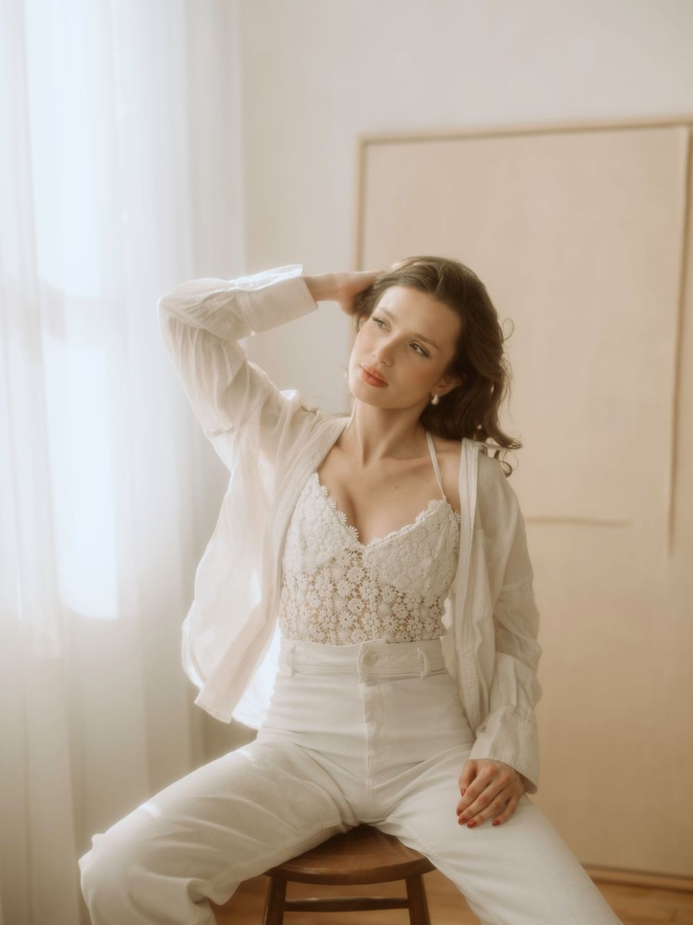Artistic portrait shows someone posing in a white shirt and pants on a wooden chair in a minimalist interior setting.