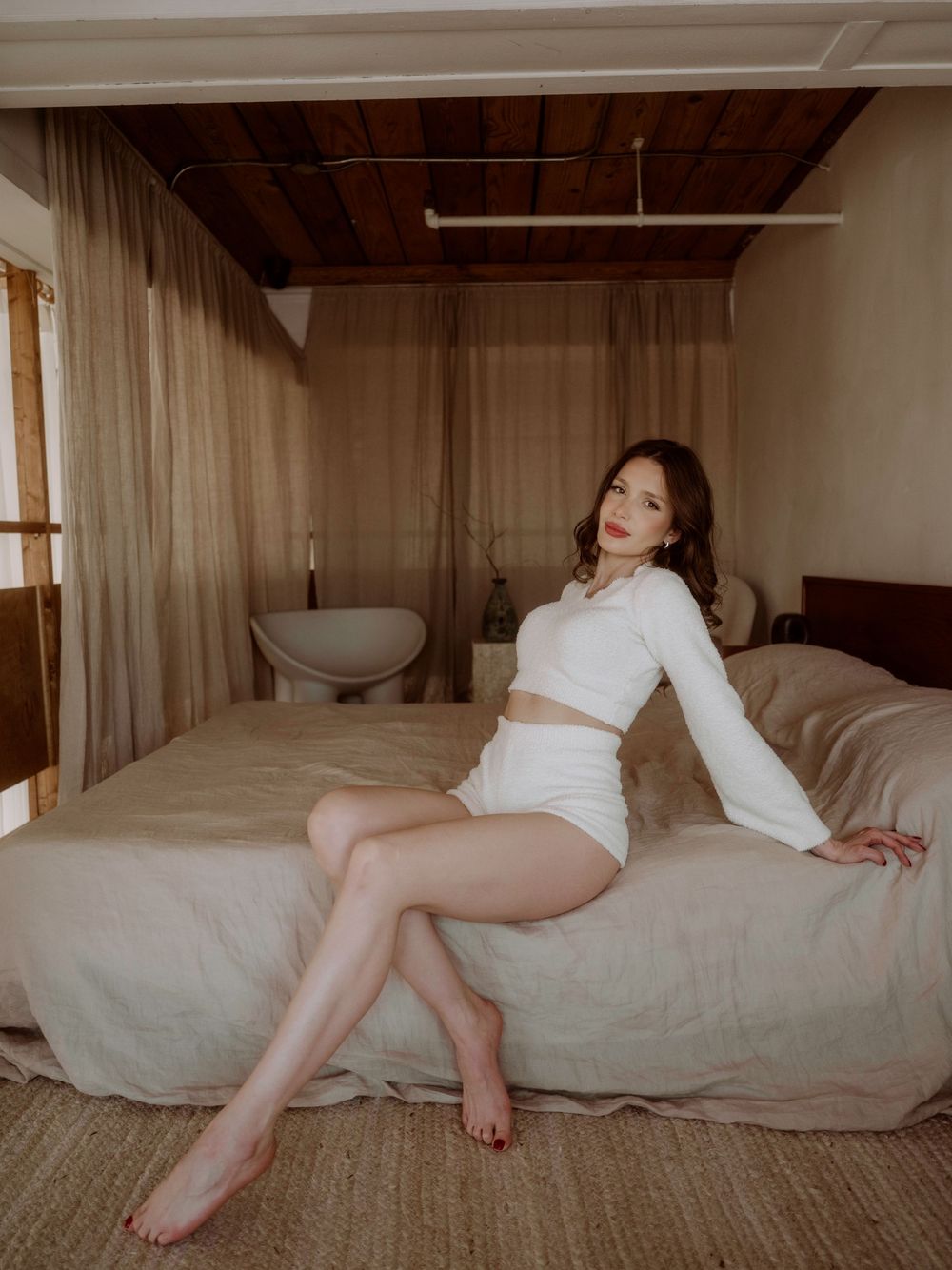 Woman in white crop top and shorts lounging on bed with natural lighting in minimalist bedroom setting.