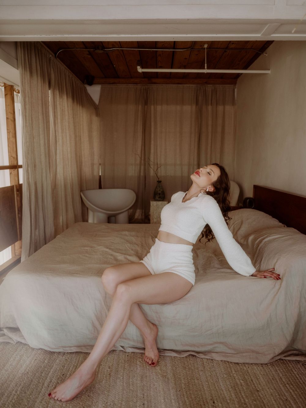 Figure in white loungewear relaxes on beige bed in minimal bedroom with sheer curtains and natural lighting.