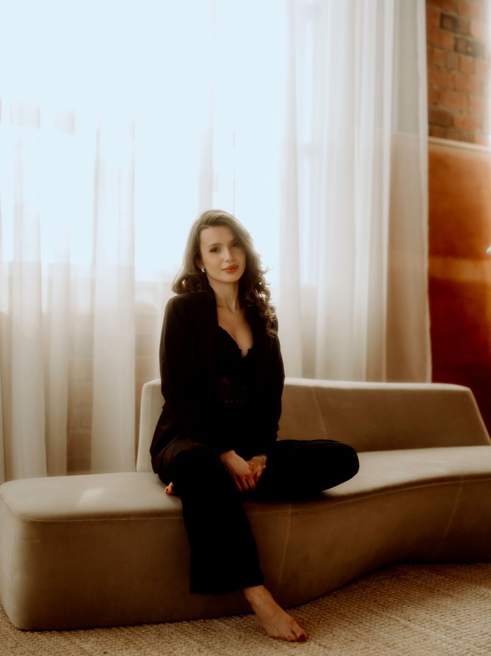 Figure in black outfit sits barefoot on a beige couch by sheer white curtains in warm, softly lit setting.