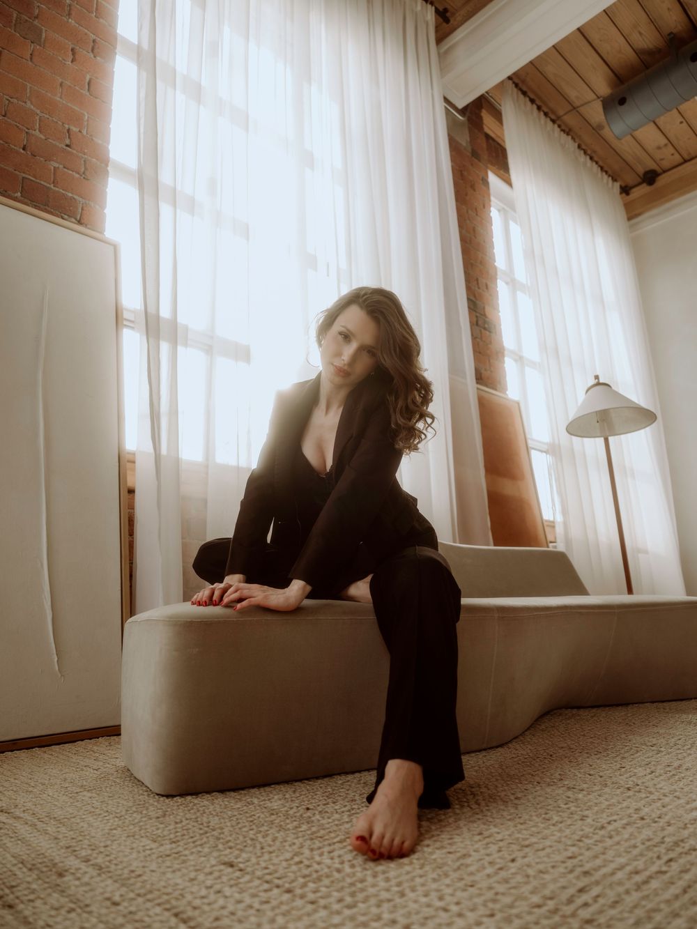 A moody portrait in a sunlit loft space with exposed brick and white curtains showing a figure in black on a beige sofa.