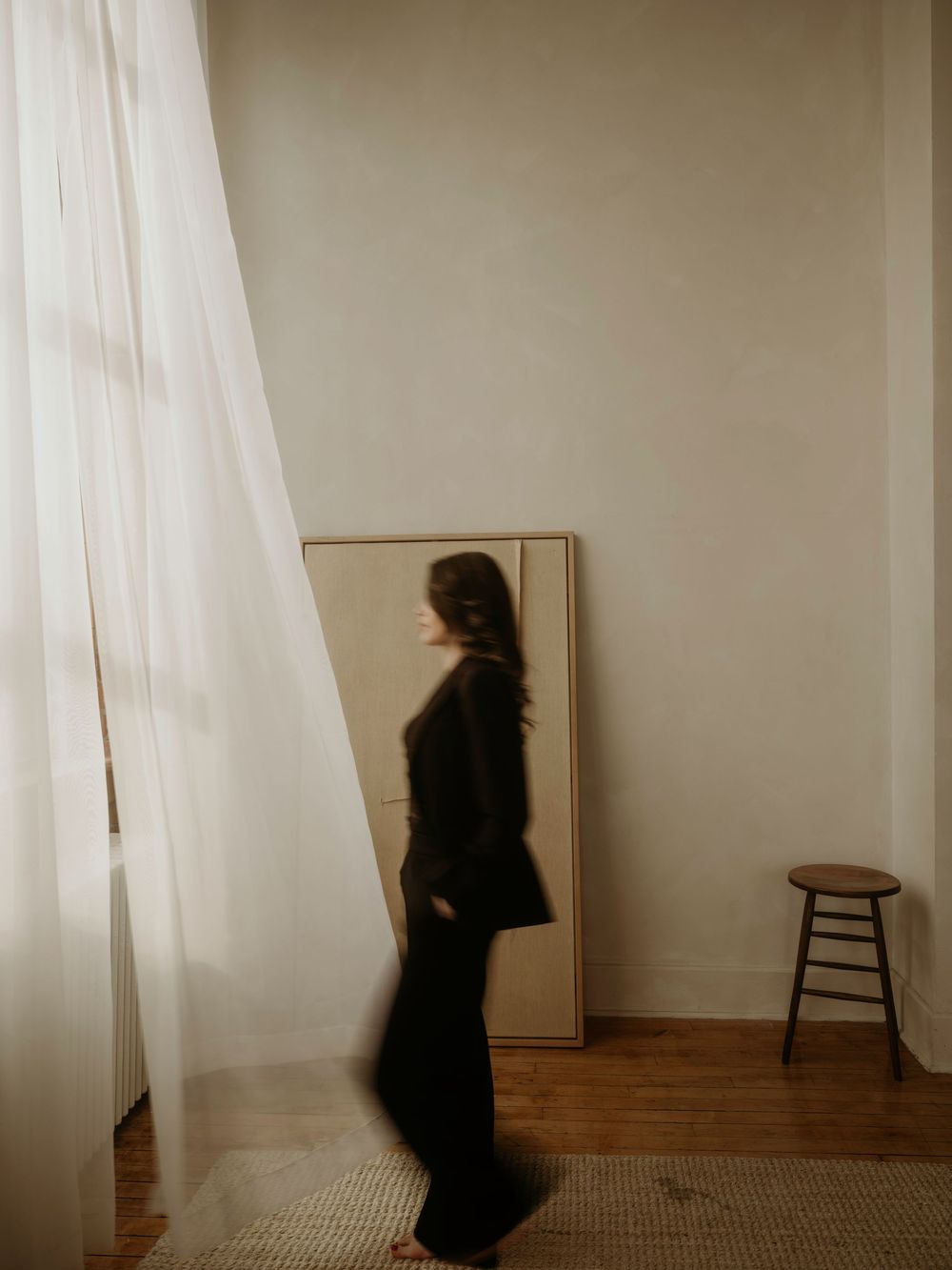 Blurry figure in black stands near a full-length mirror and wooden stool in a minimalist room with sheer curtains.