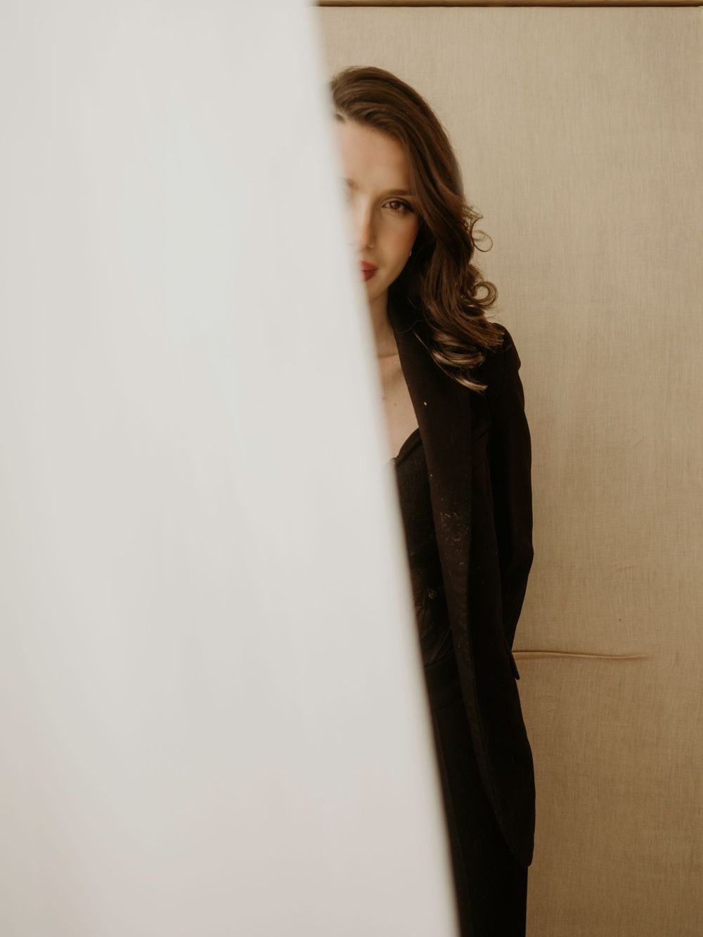 Artistic portrait of someone partially hidden behind a light colored wall in moody lighting wearing dark clothing.