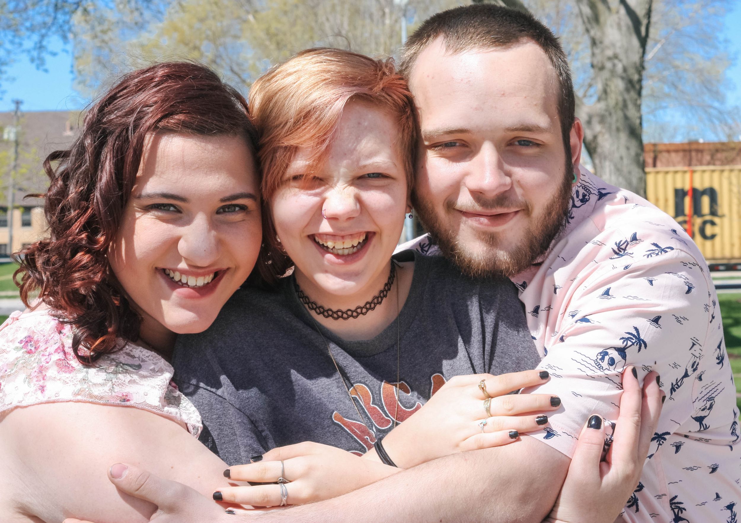 Three friends share a joyful embrace outdoors on a sunny day with trees and buildings in the background.