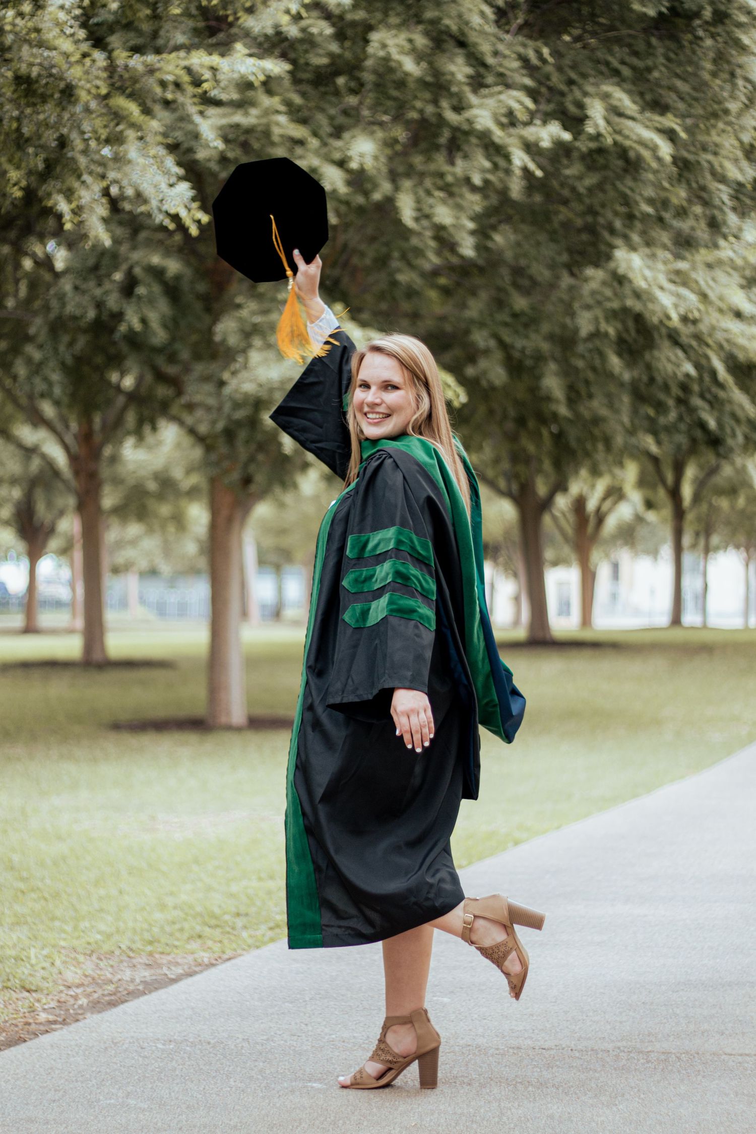Graduate in black and green academic regalia poses playfully on campus pathway while holding cap aloft.