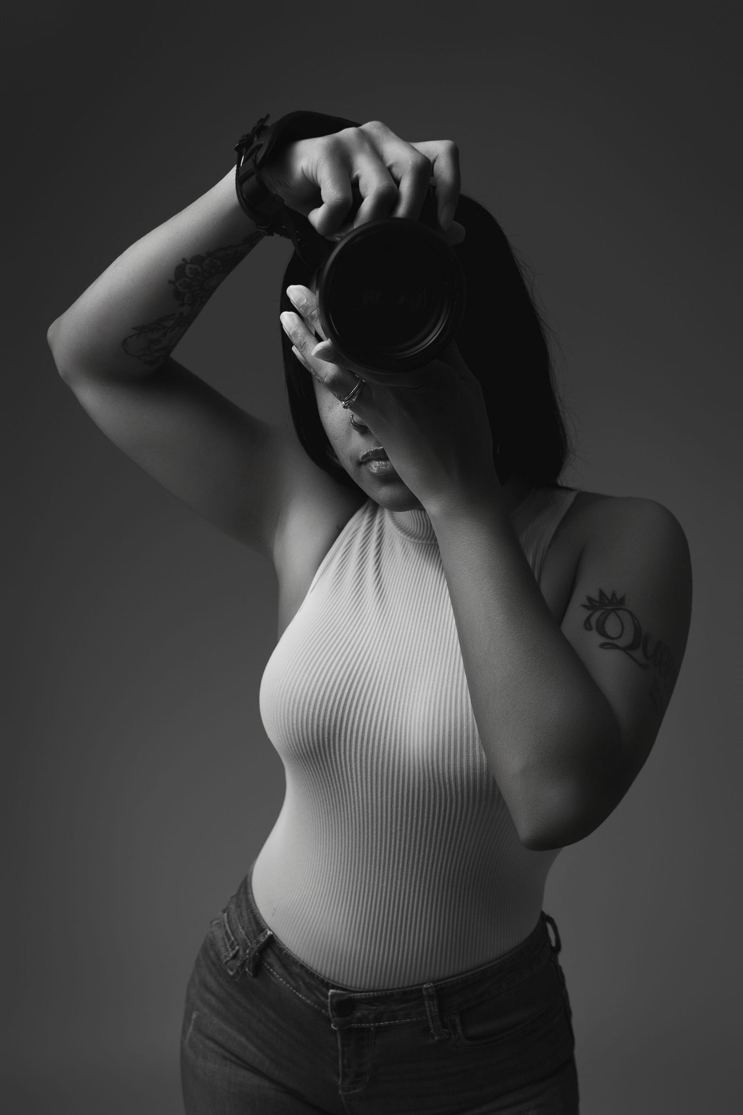 A photographer holding a camera in a white ribbed tank top and jeans poses in dramatic black and white lighting.