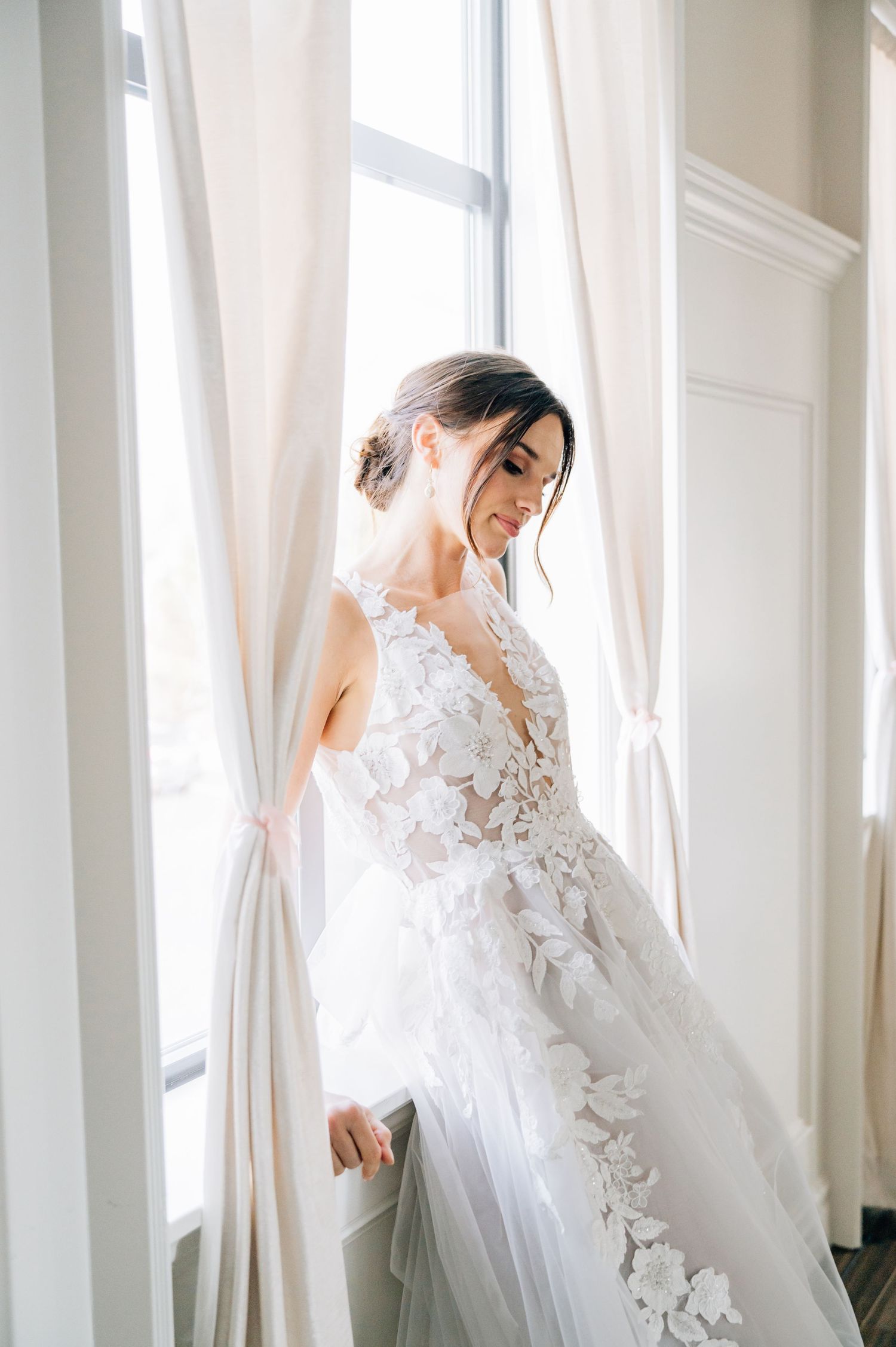 Ethereal wedding gown with delicate floral appliques and sheer details captured in soft natural light by a window.